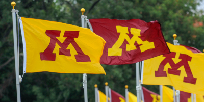 U of M maroon and gold flags wave in the wind against a backdrop of green trees